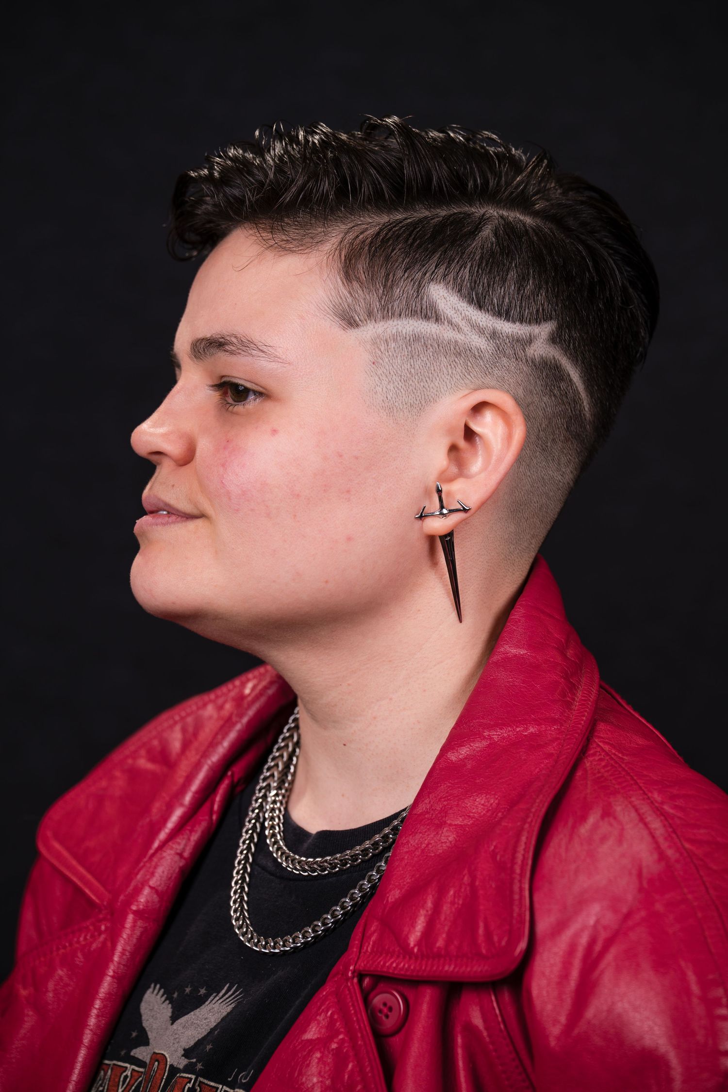 Side profile portraits in red leather jacket against dark background showing short hairstyle and silver chain necklace.