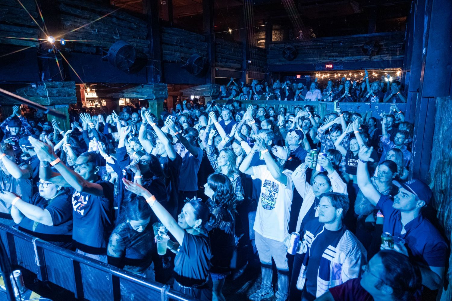Concert crowd with raised arms illuminated in blue lighting during a live performance at an indoor venue.