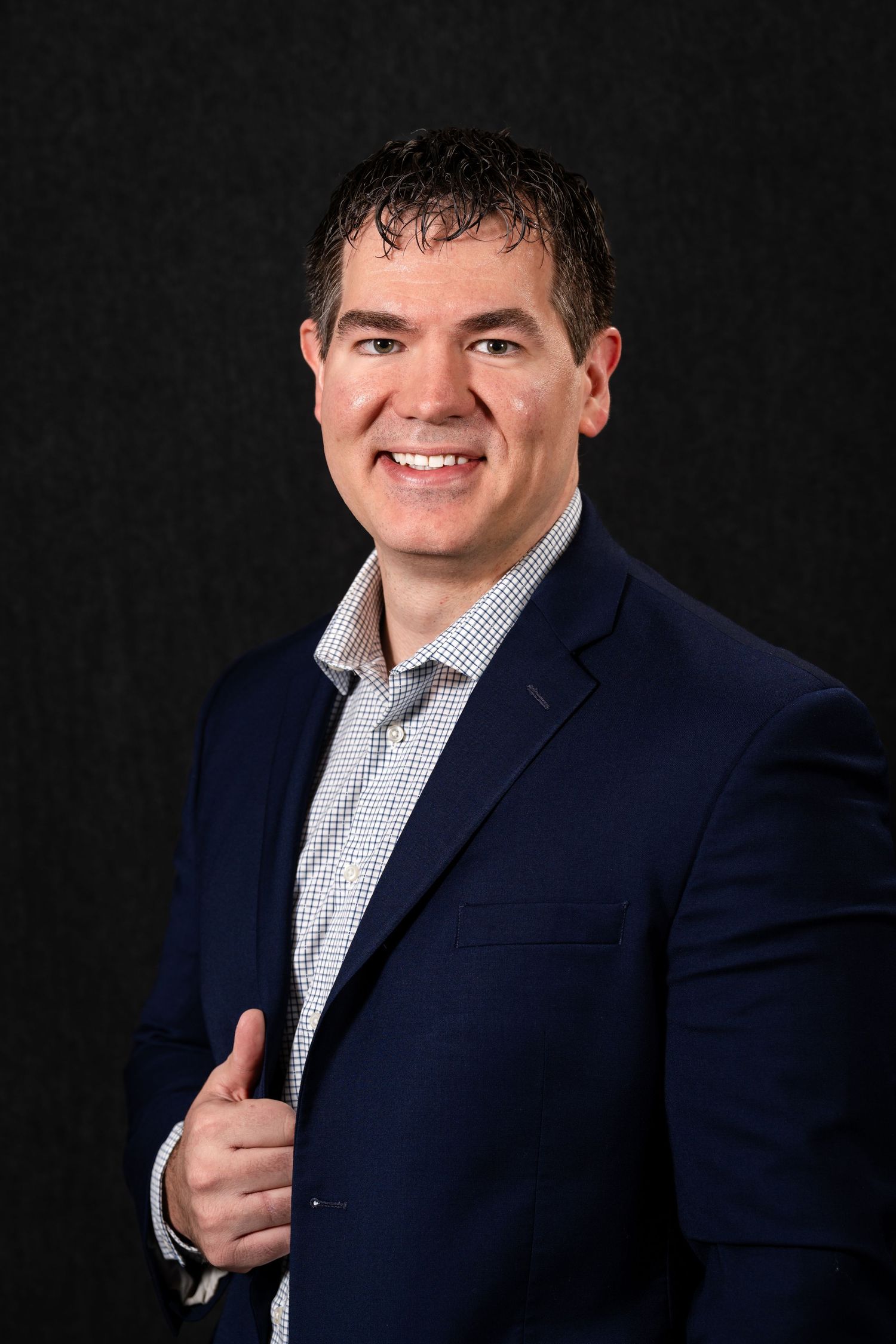 Professional headshot portrait of someone in a navy suit jacket and checkered dress shirt against a dark background.