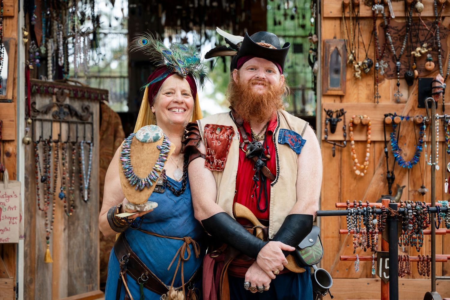 Two people in renaissance festival costumes smile while sitting together against a rustic wooden backdrop with hanging decor.