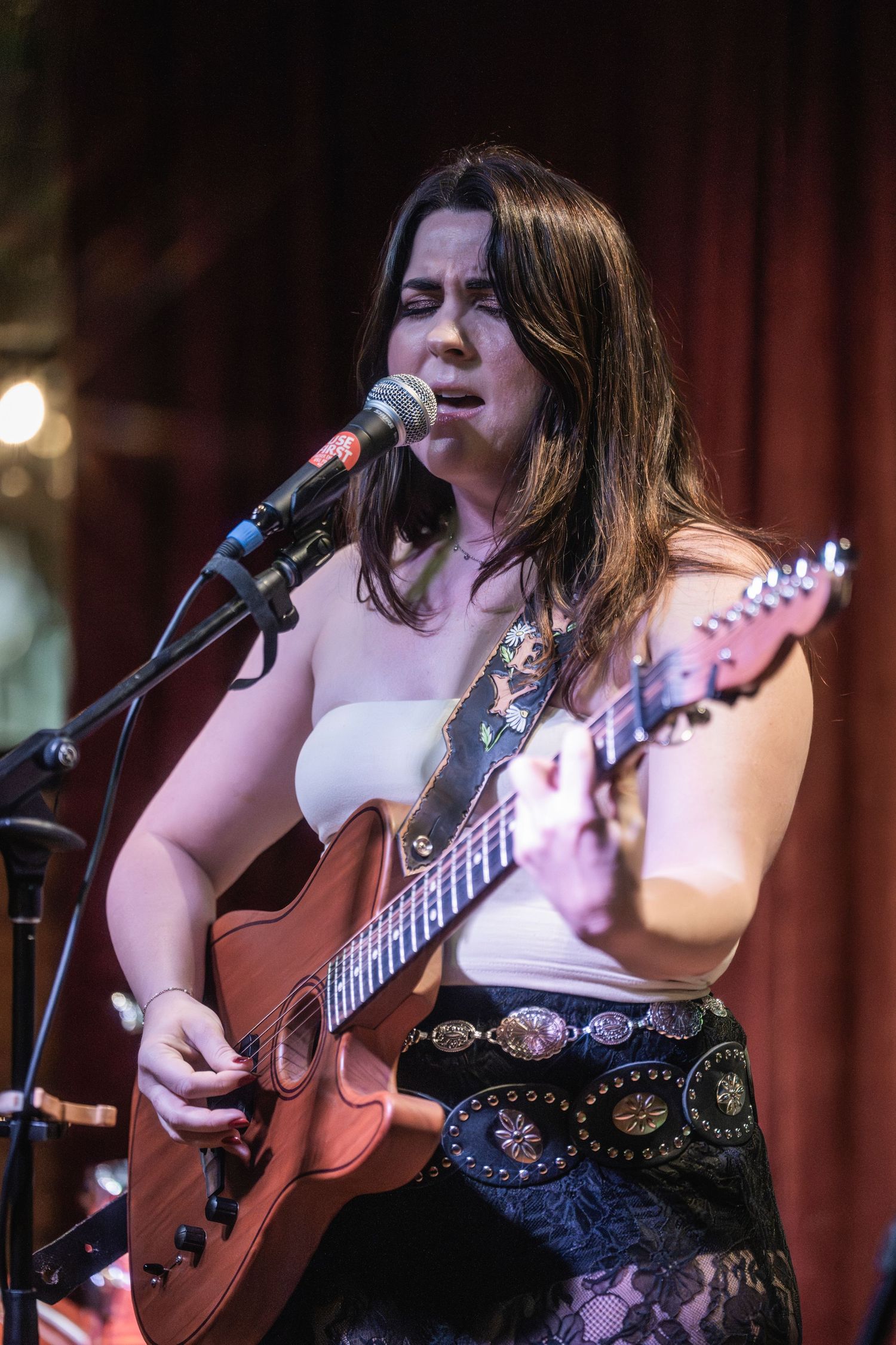 A musician performs with an acoustic guitar on stage in a dimly lit venue wearing a black lace top.