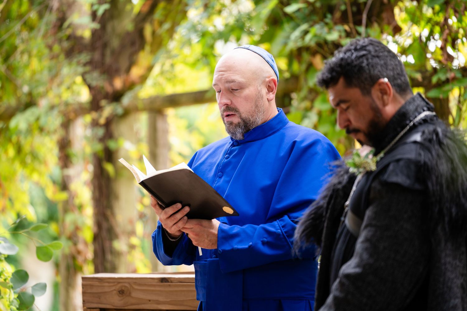 Person in blue jacket using digital tablet while sitting outdoors with companion in wooded setting.