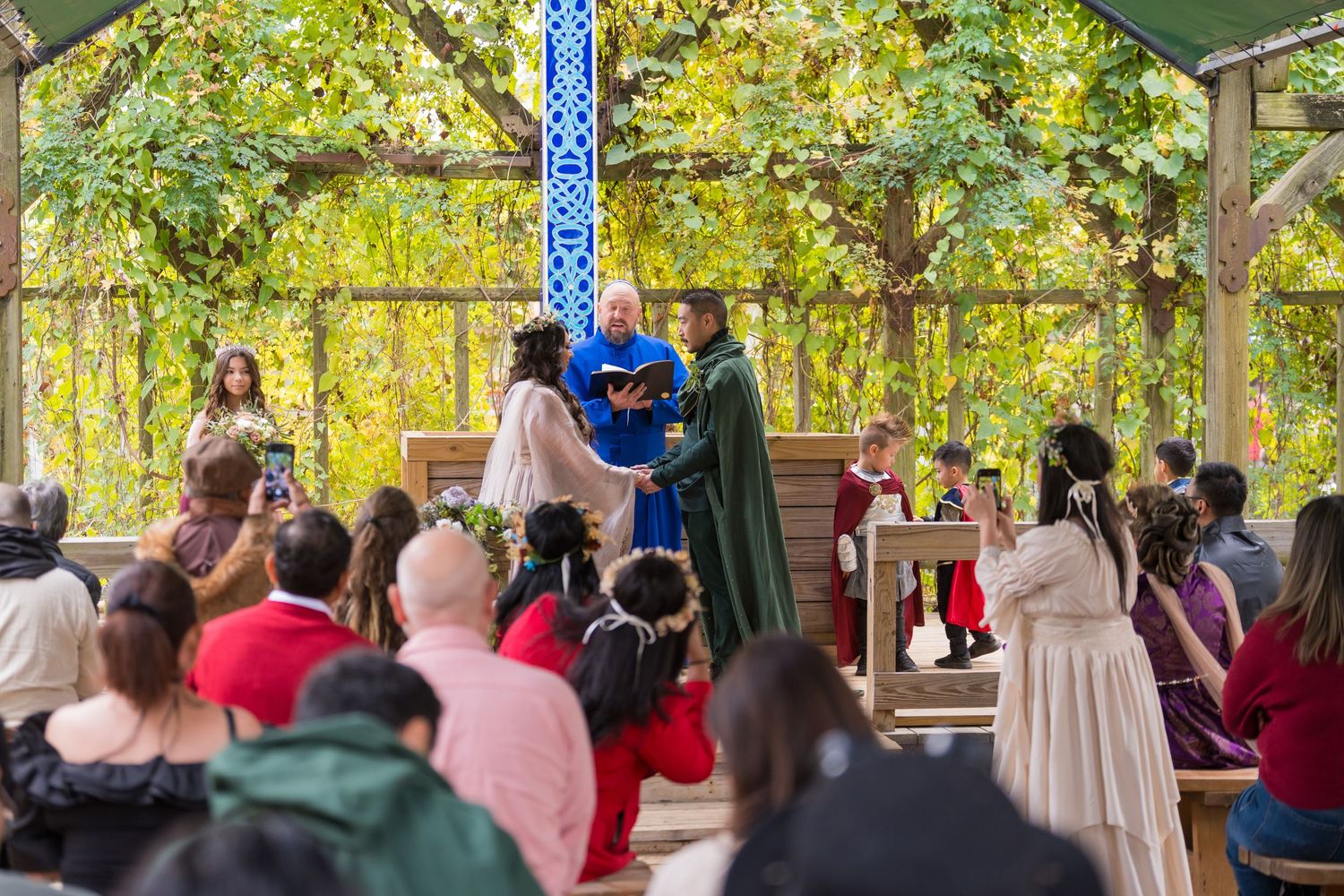 A wedding ceremony takes place under a wooden pergola surrounded by autumn foliage and seated guests.