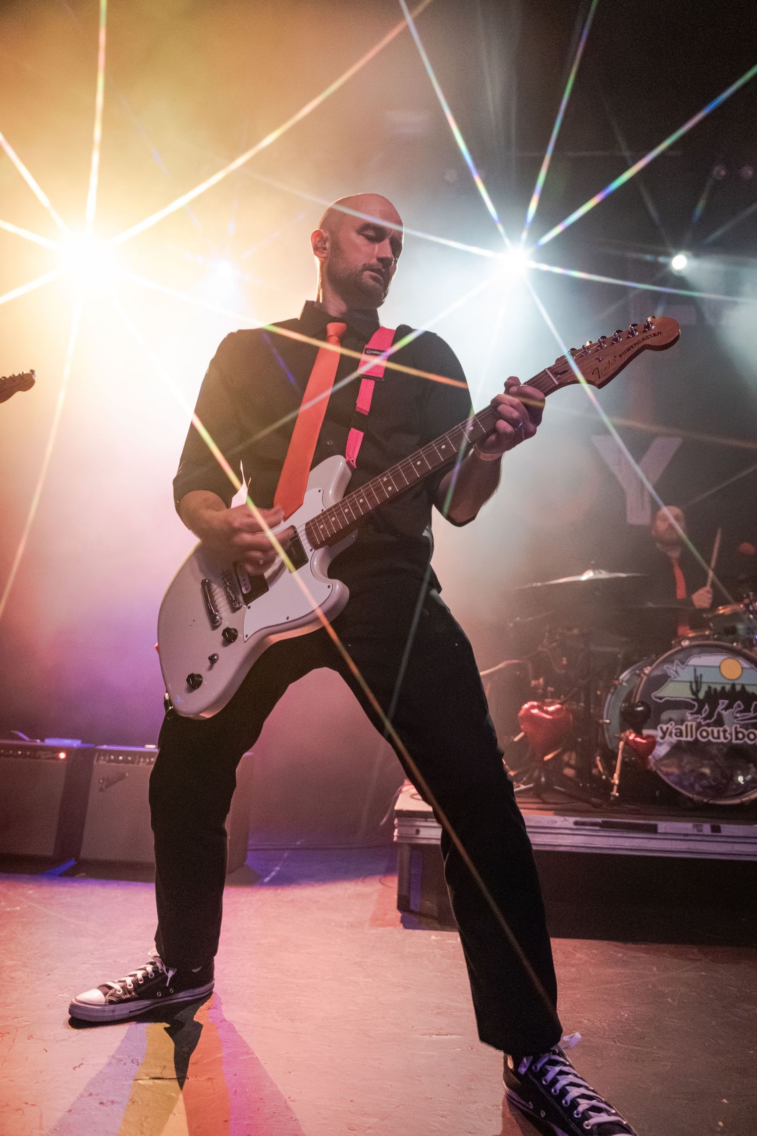 Bald guitarist performs on stage with white electric guitar, dramatic laser lights and drum kit visible behind him.