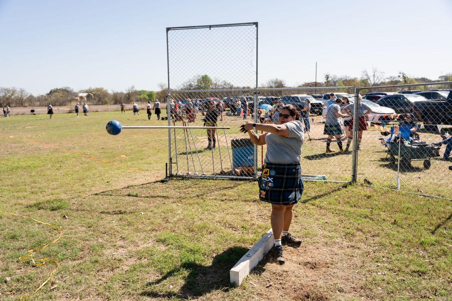 Athlete in kilt competes in Highland Games hammer throw on grassy field with spectators watching in background.