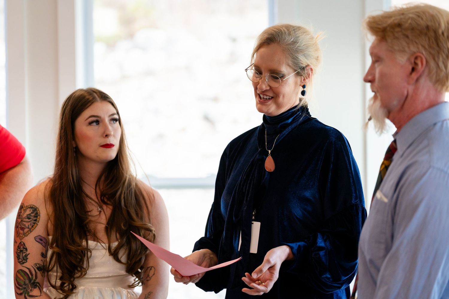 Woman in white dress with tattoos speaks with officiant in blue at an indoor wedding ceremony.