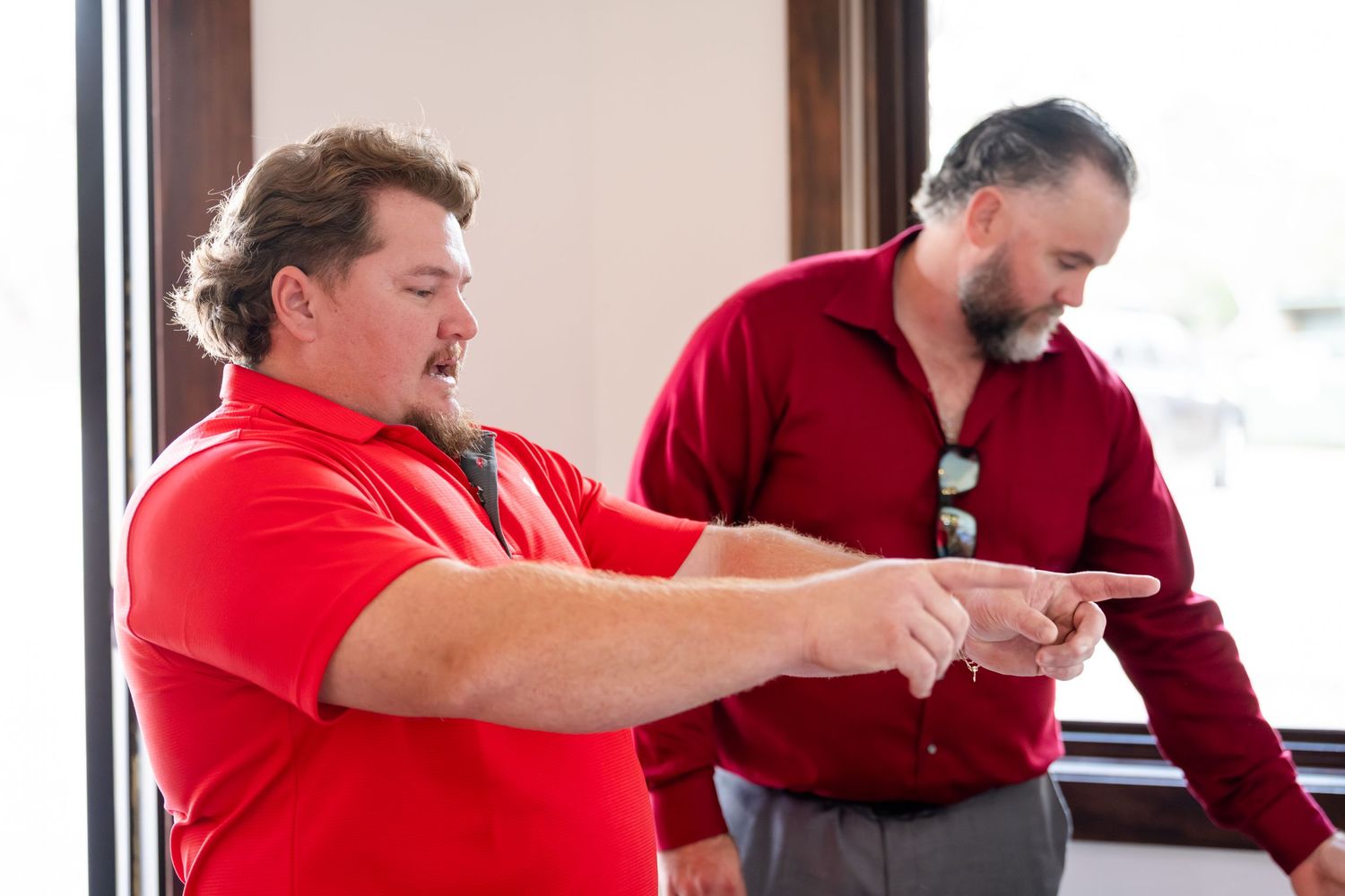 Two men in red shirts, one pointing finger, engaged in discussion near a bright window.