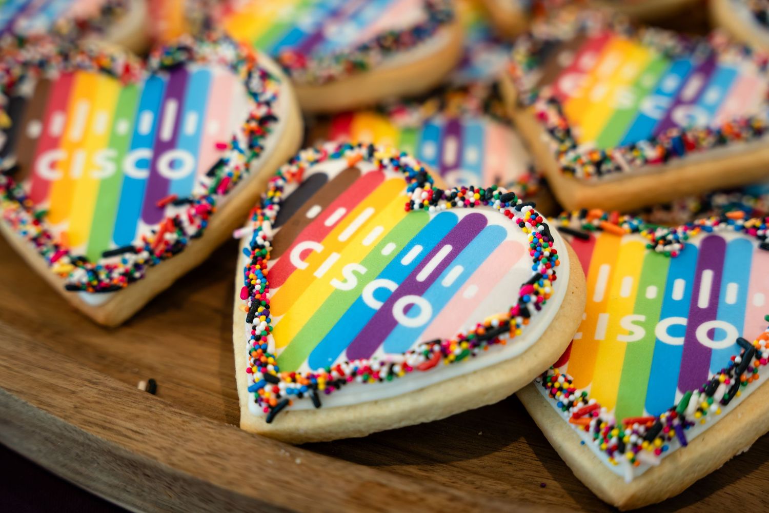 Heart-shaped Cisco logo sugar cookies with rainbow stripes and colorful sprinkles on a wooden tray.