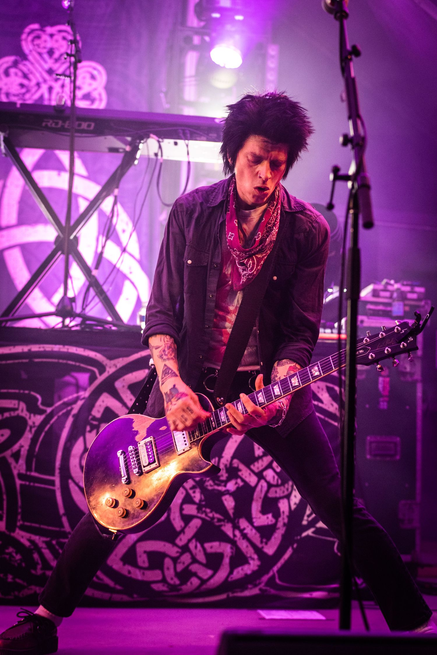 Guitarist performs on stage bathed in purple lighting against graffiti backdrop during energetic rock concert performance.