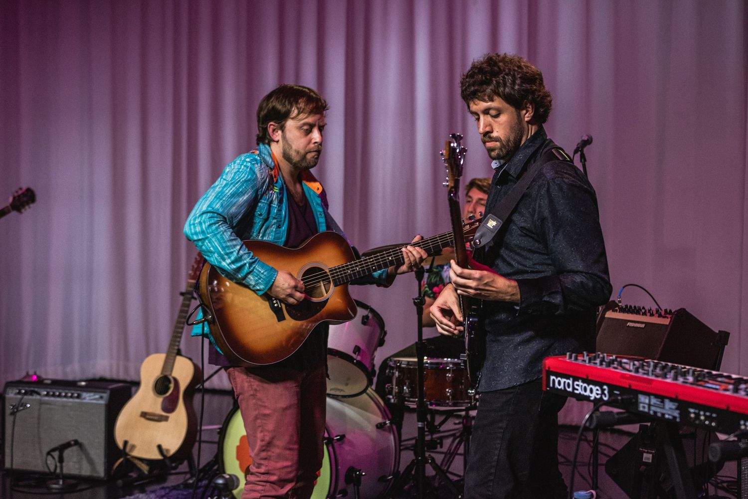 Musicians perform on stage with acoustic guitars and instruments against a purple-lit backdrop during a live concert.