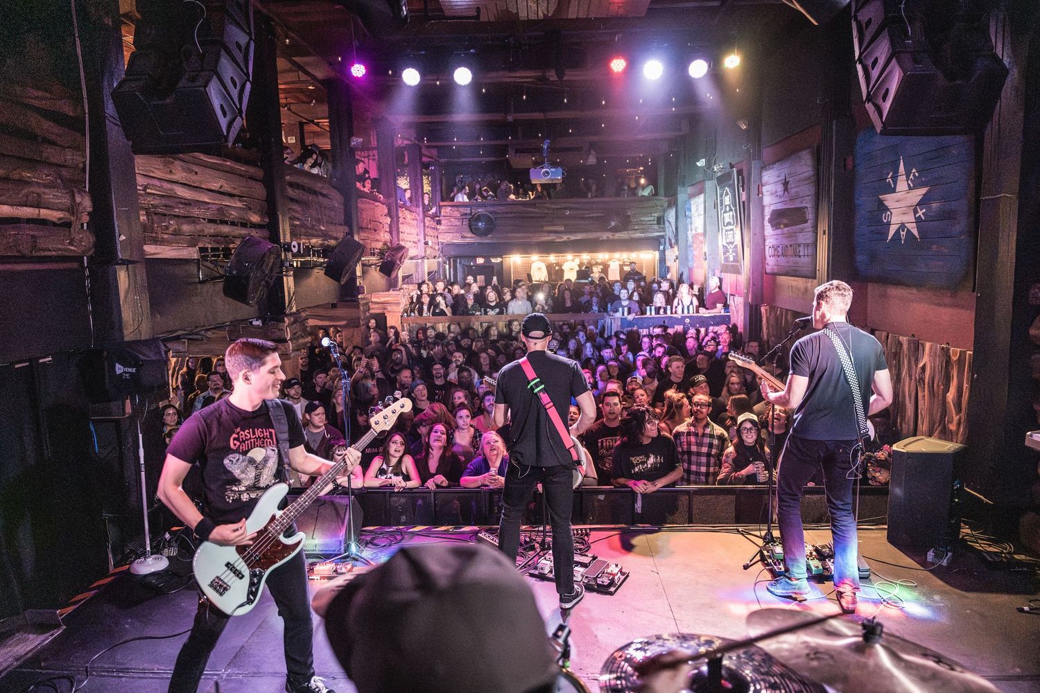 A rock band performs on stage at a crowded music venue with purple lighting and American flags on the walls.