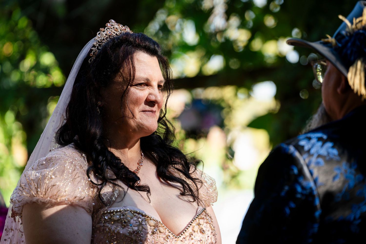A bride in a blush pink lace wedding dress smiles outdoors on a sunny day during a ceremony.