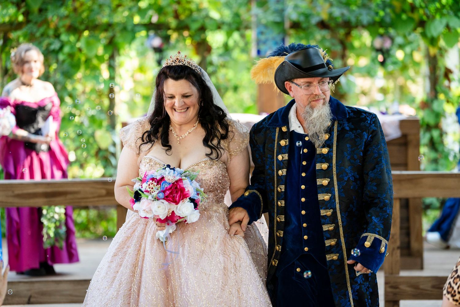 A couple in formal wedding attire walks together in a garden setting with bright greenery in the background.