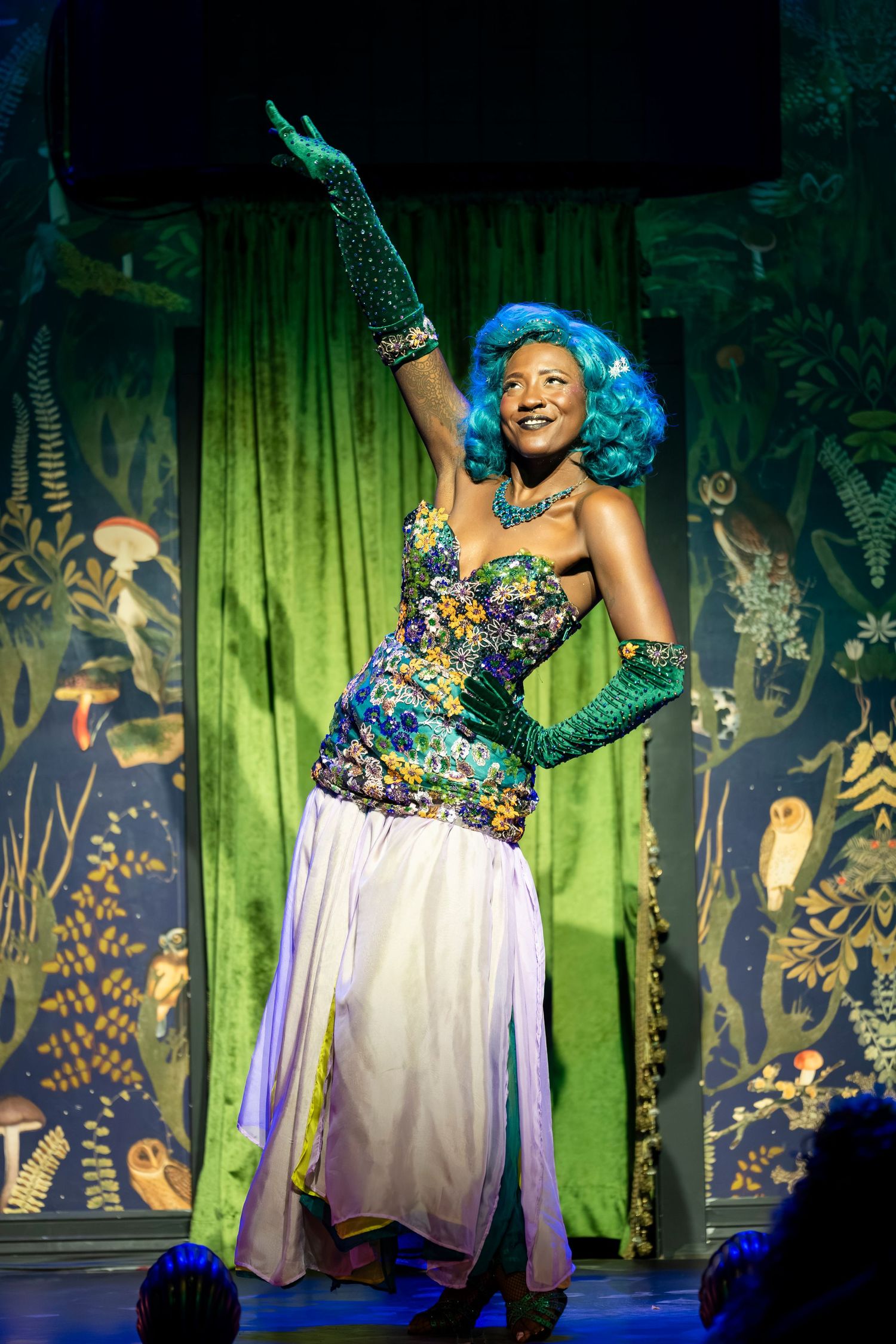 A performer in a sparkly corset, blue wig and long gloves strikes a dramatic pose on stage with green curtains behind.