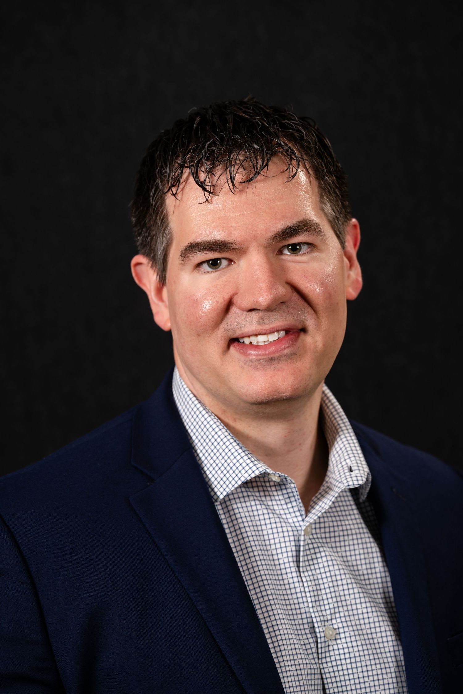 Professional headshot portrait of a person in a navy blazer and checkered dress shirt against a dark background.