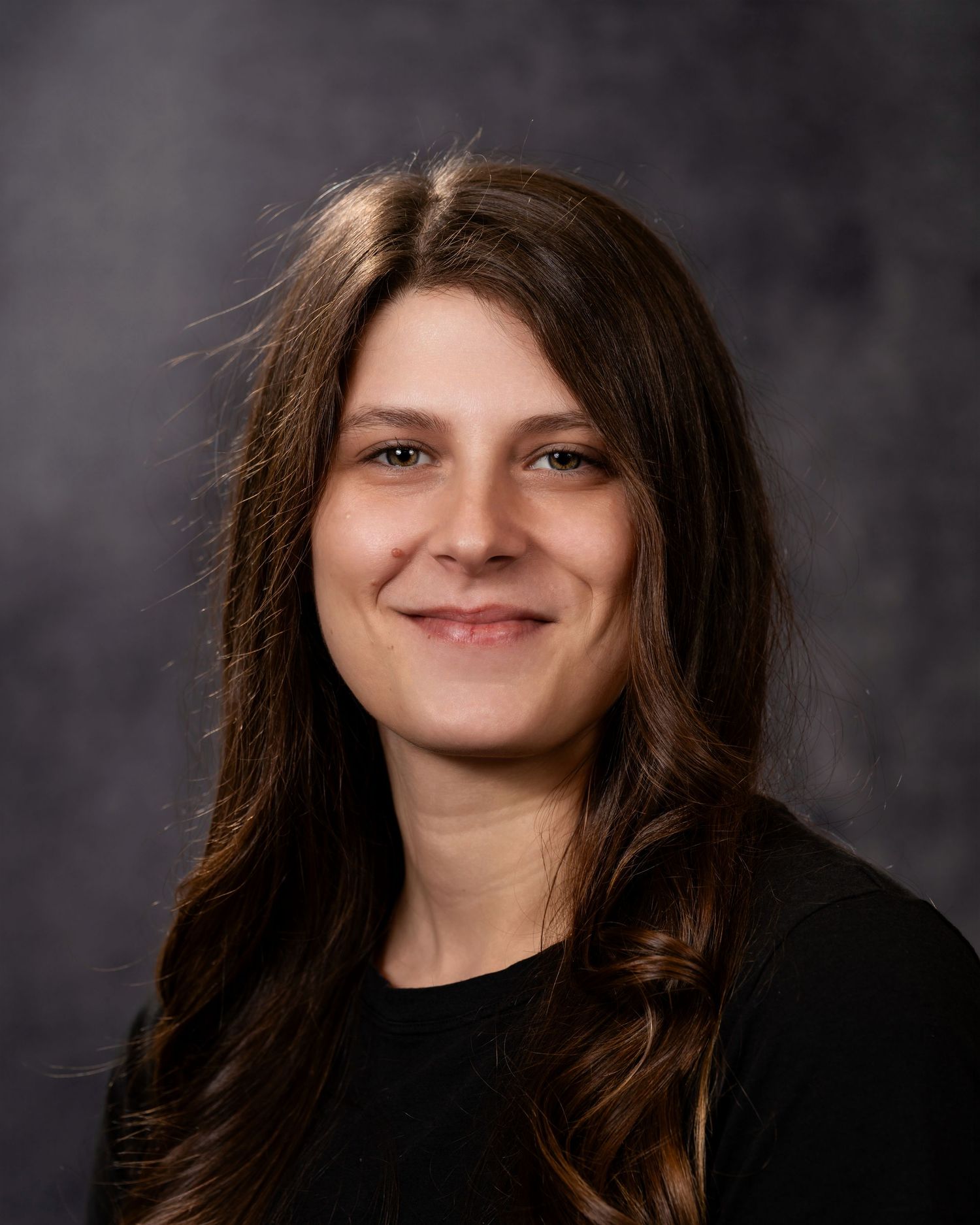 Professional headshot portrait against a dark gray background wearing a black top with long brown hair.