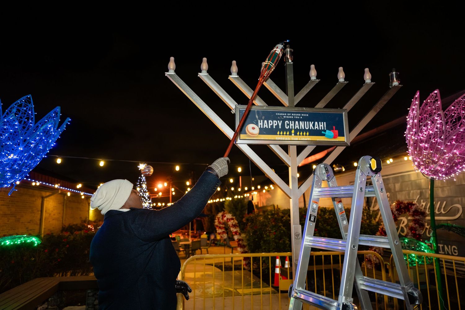 A large menorah illuminated at night with blue and pink lit decorations visible in an outdoor holiday display.