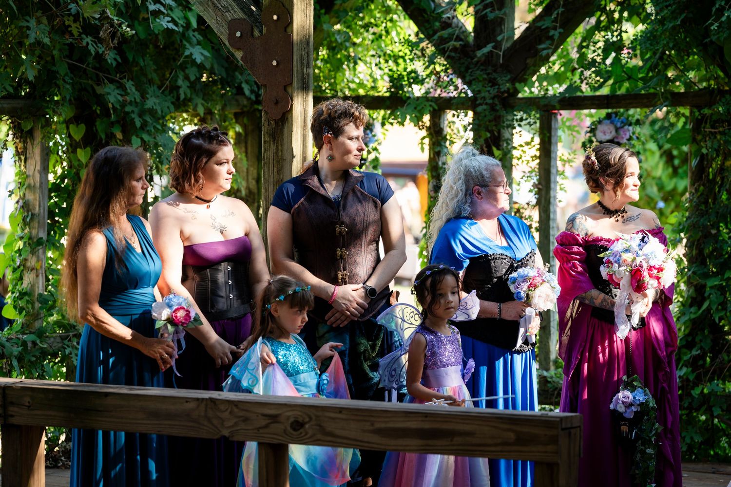 A group of people gather under a wooden pergola surrounded by lush greenery on a sunny day.