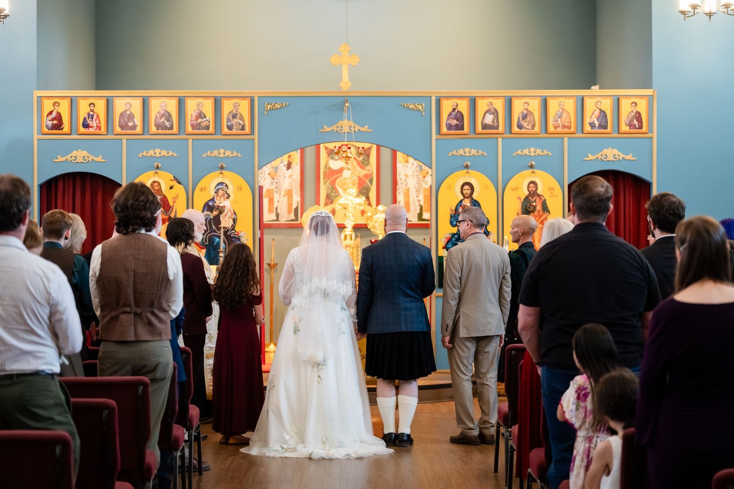 Wedding ceremony taking place in an Orthodox church with gold icons and blue walls in the background.
