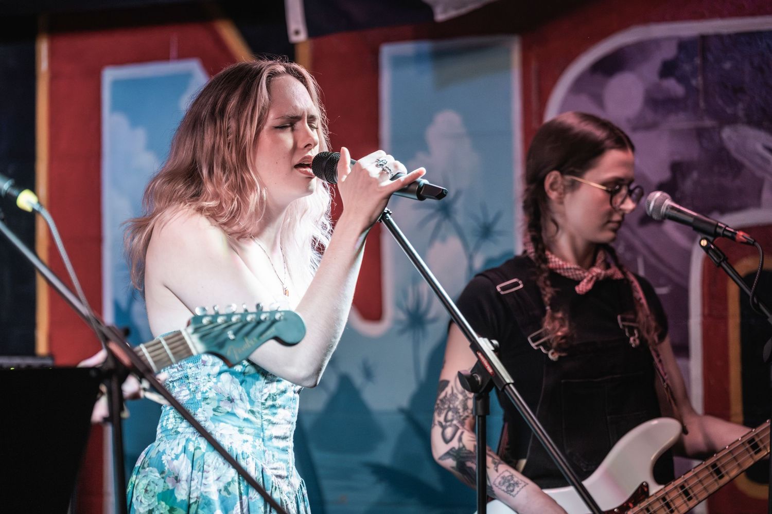 Musicians perform on stage with microphones and instruments against a red backdrop during a live concert.