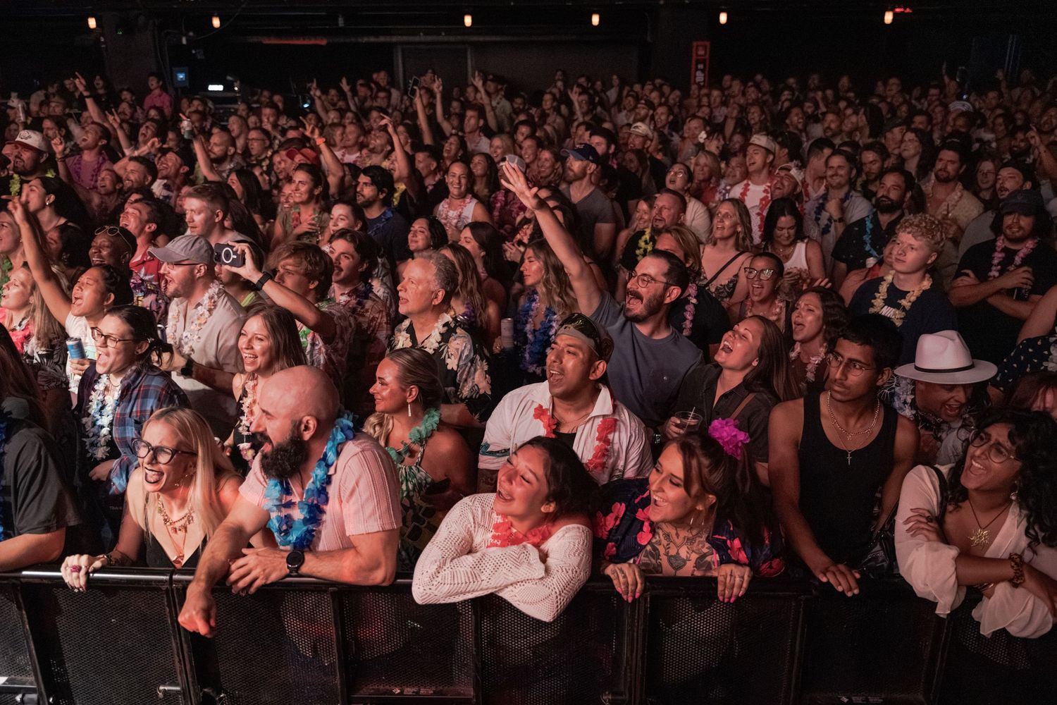 Large diverse crowd of concert fans enjoying a live music event indoors, some raising hands and cheering.