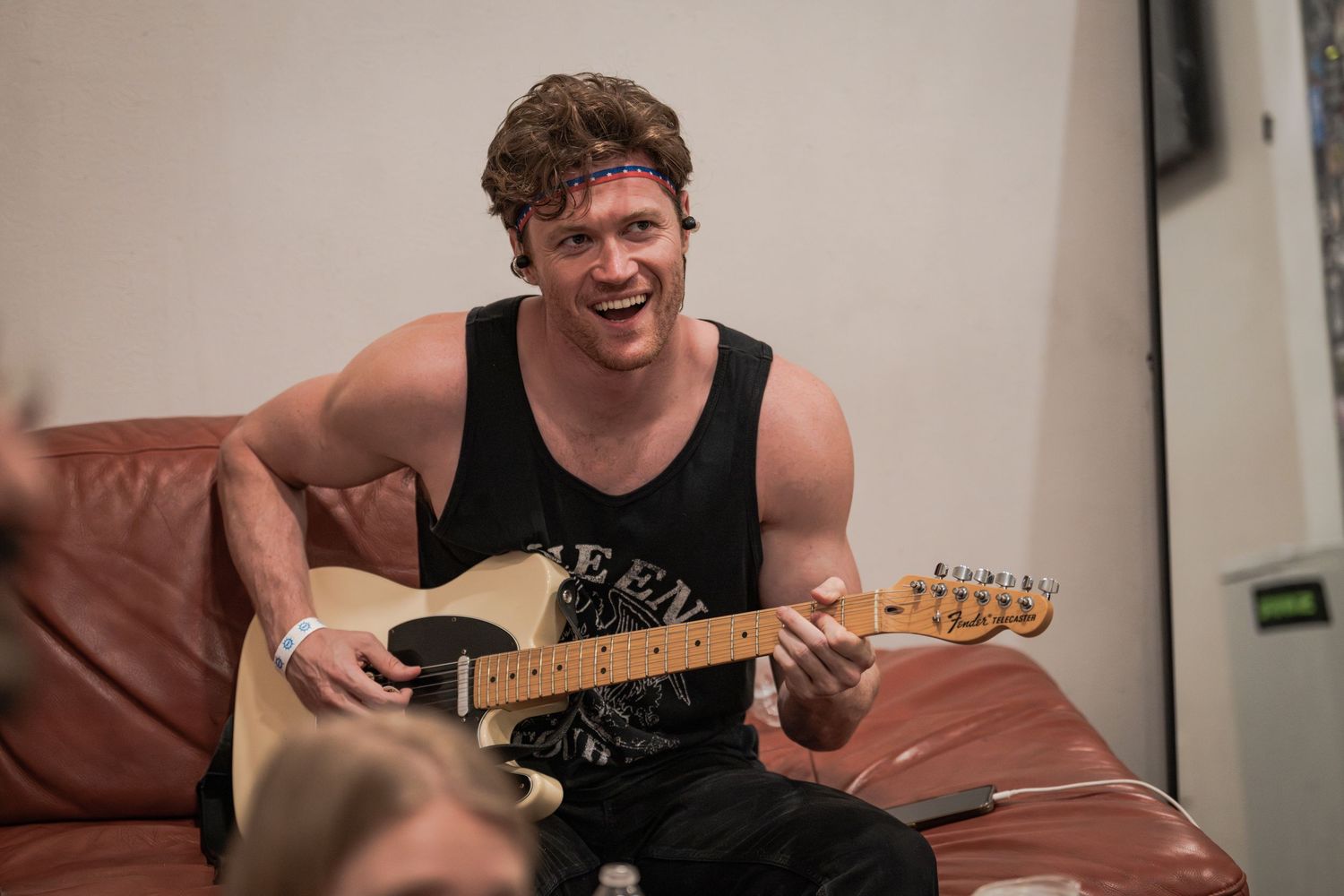 Smiling young man playing electric guitar on brown leather couch wearing black tank top and purple headband.