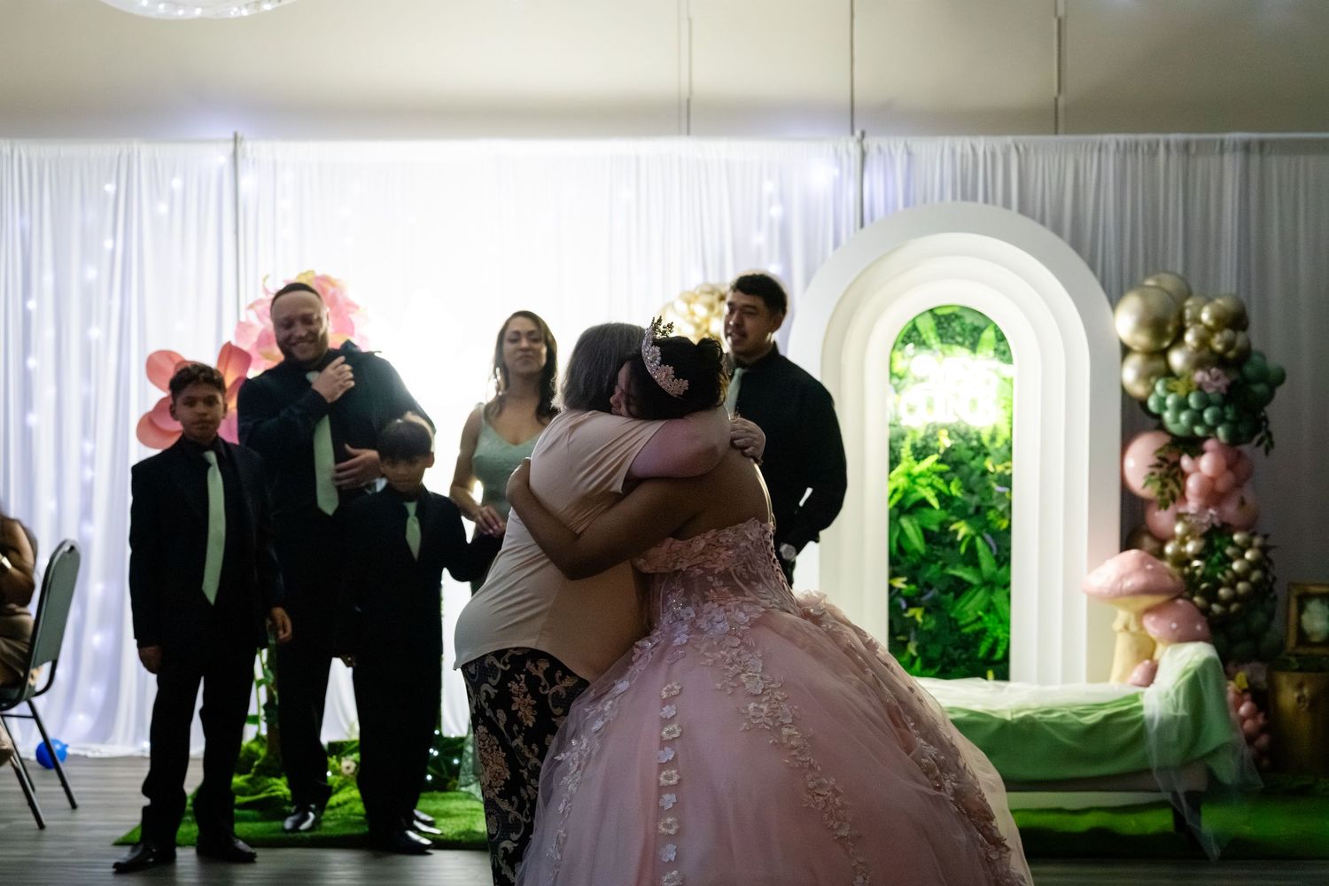 A girl in a pink quinceañera gown shares a heartfelt hug with a guest at her celebration party.