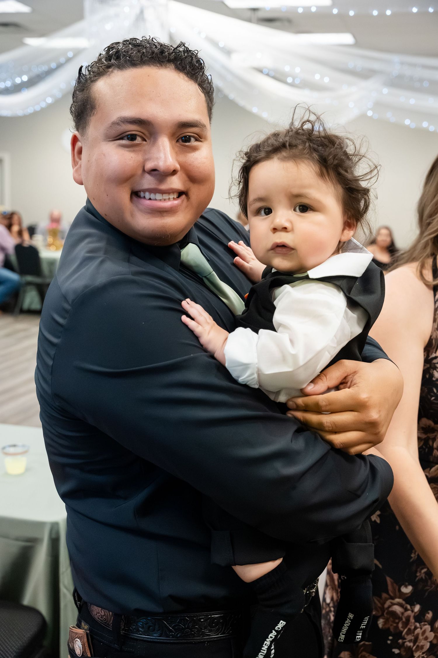 Man in black suit holding a dressed-up baby at a formal event celebration.
