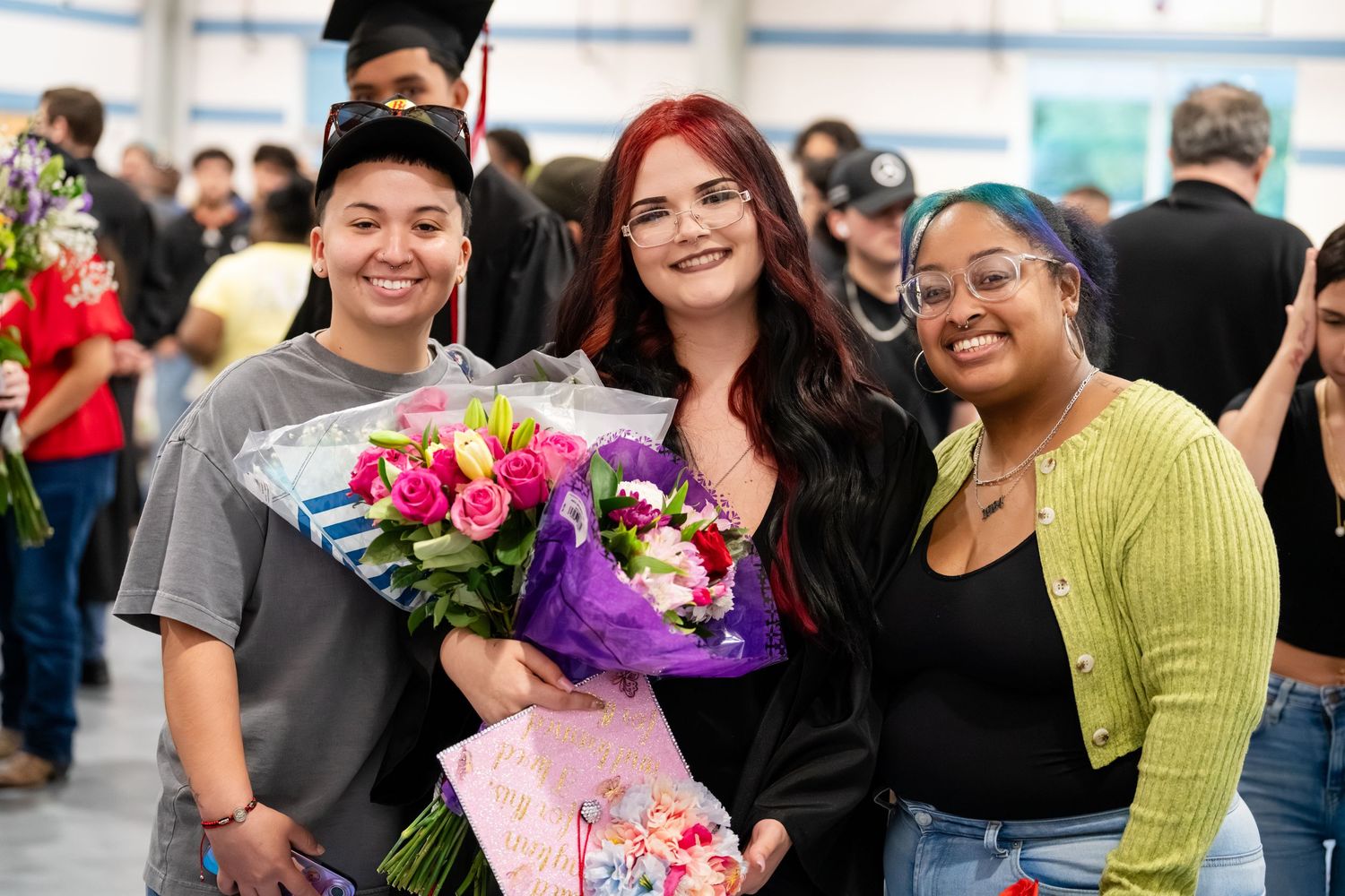 Three smiling young women pose together at a graduation celebration, holding colorful flower bouquets.