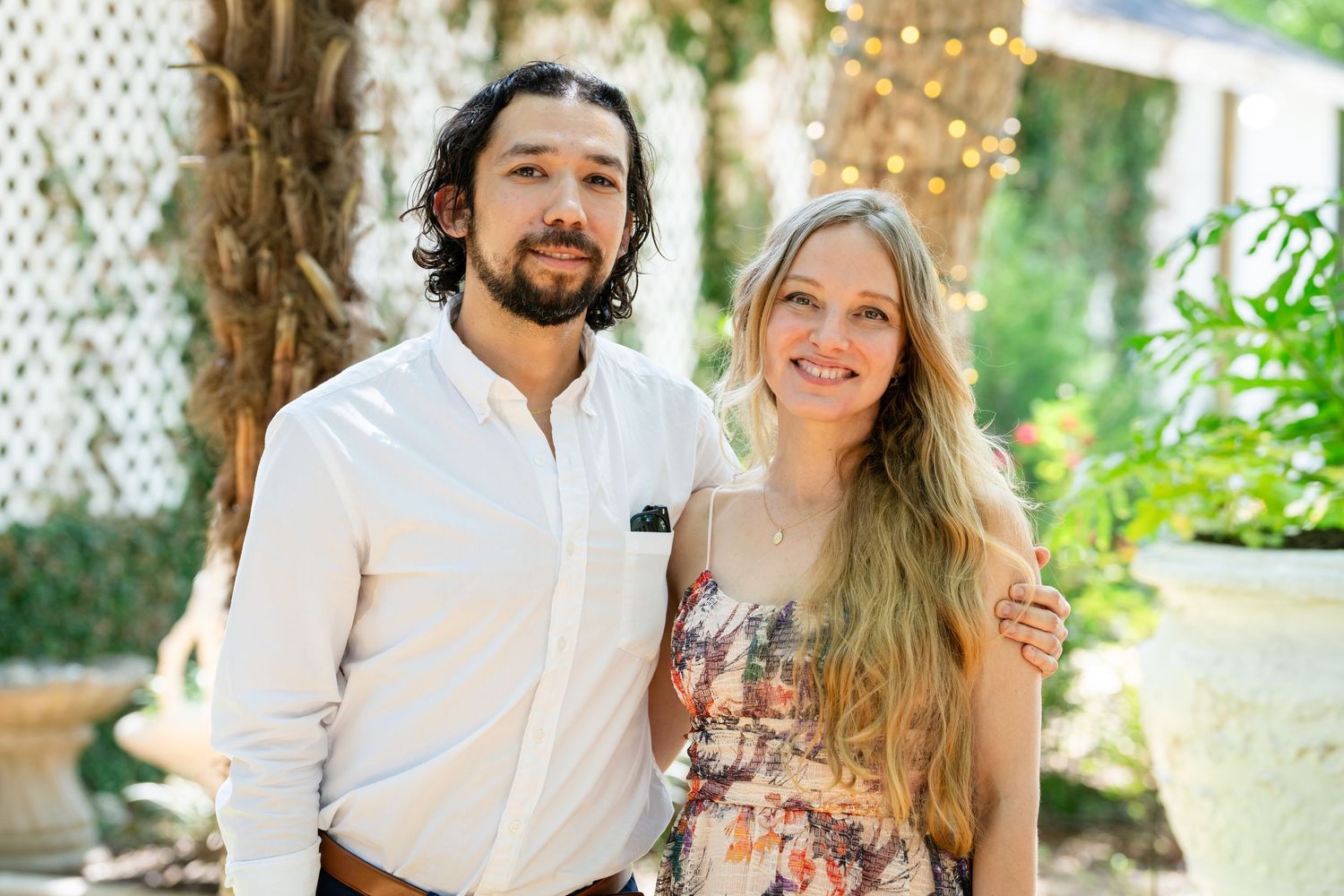 A smiling couple poses together outdoors in a garden setting, man in white shirt with woman in floral dress.