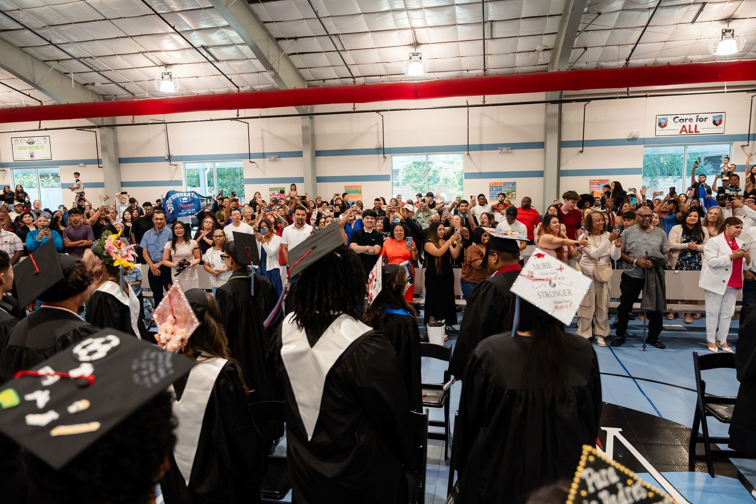 Graduation ceremony in a gymnasium with graduates in caps and gowns facing a large crowd of cheering attendees.