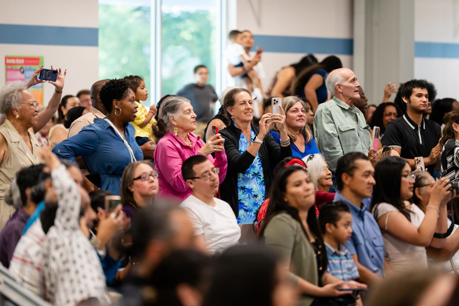 Diverse crowd of people standing and taking photos at an indoor community event or gathering.
