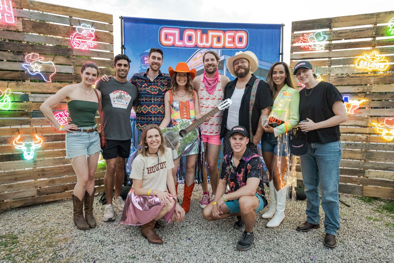 Group of friends posing together at a Glowdeo neon sign event, smiling outdoors on a gravel area.