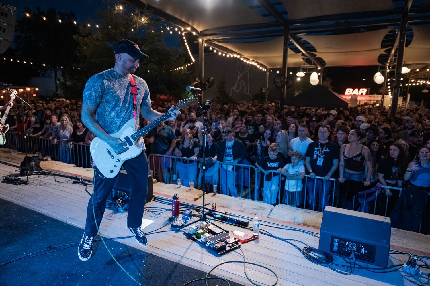 Tattooed guitarist performs on outdoor stage at night before a large crowd with string lights and a bar in the background.