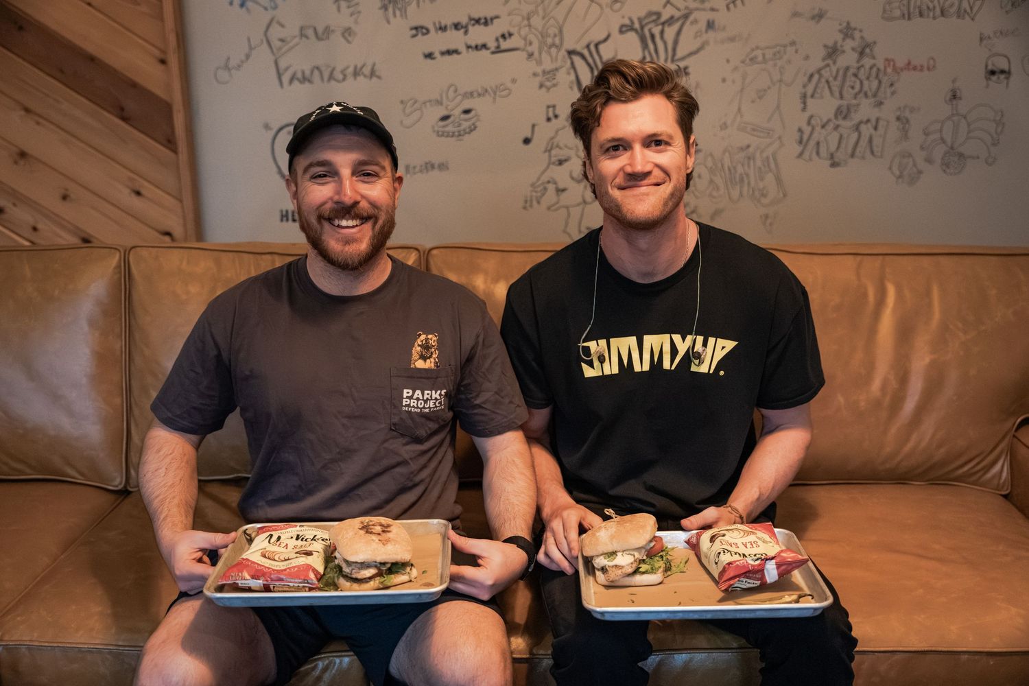 Two smiling men sitting on a couch holding trays of sandwiches, one wearing a Yummy t-shirt.