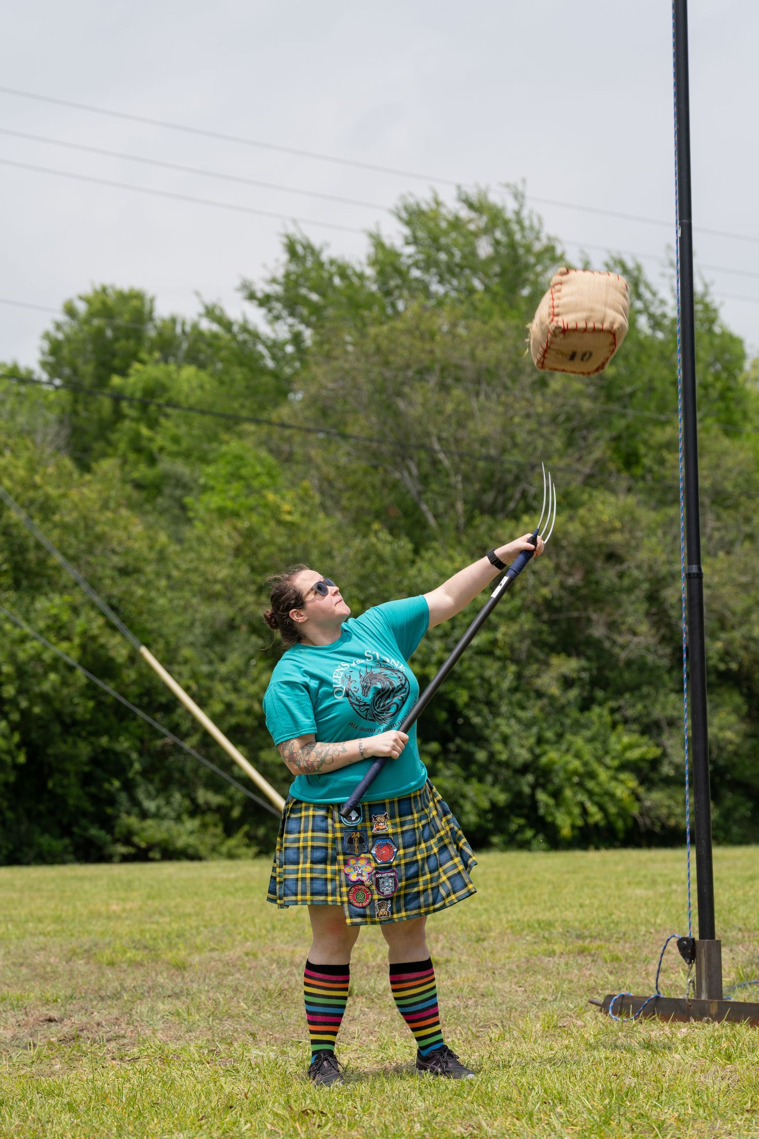 Person in kilt and teal shirt tossing a sheaf with a pitchfork at a Scottish Highland Games competition outdoors.