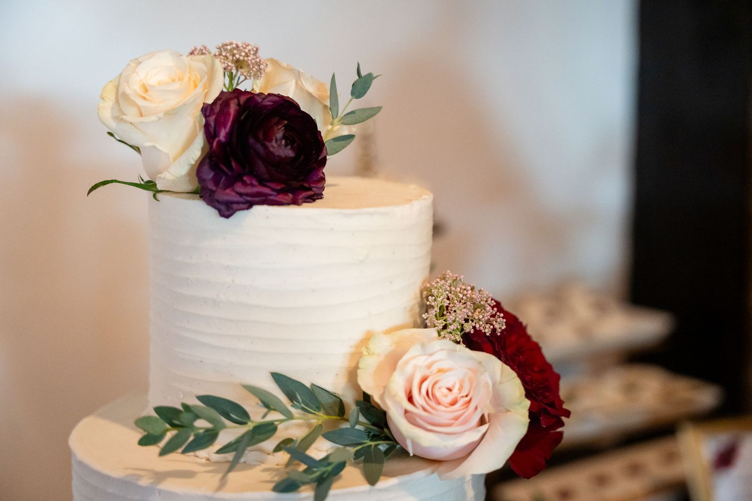 White wedding cake detail with burgundy and blush roses and greenery decorations.