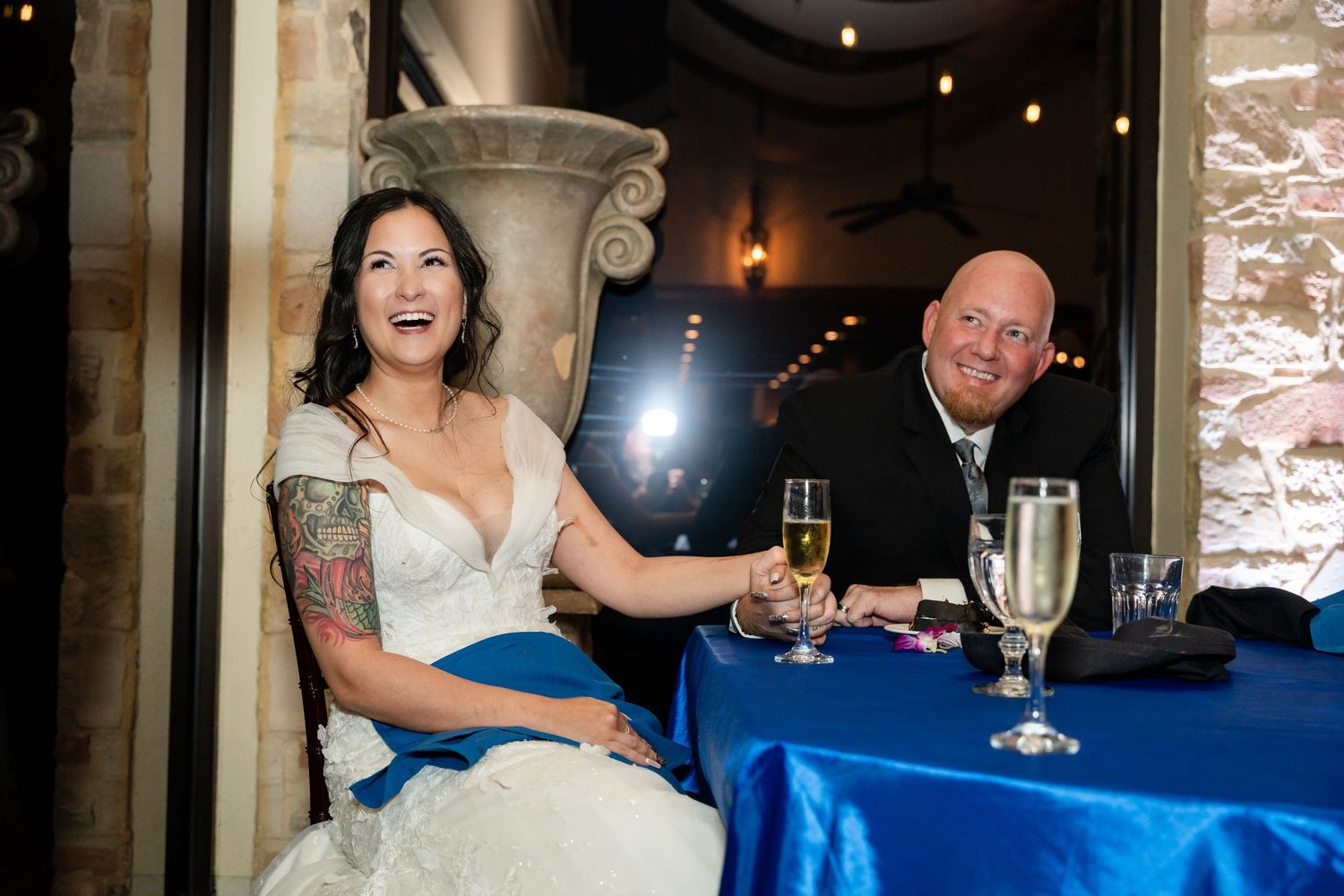Couple enjoying champagne at reception with blue tablecloth and stone architecture.