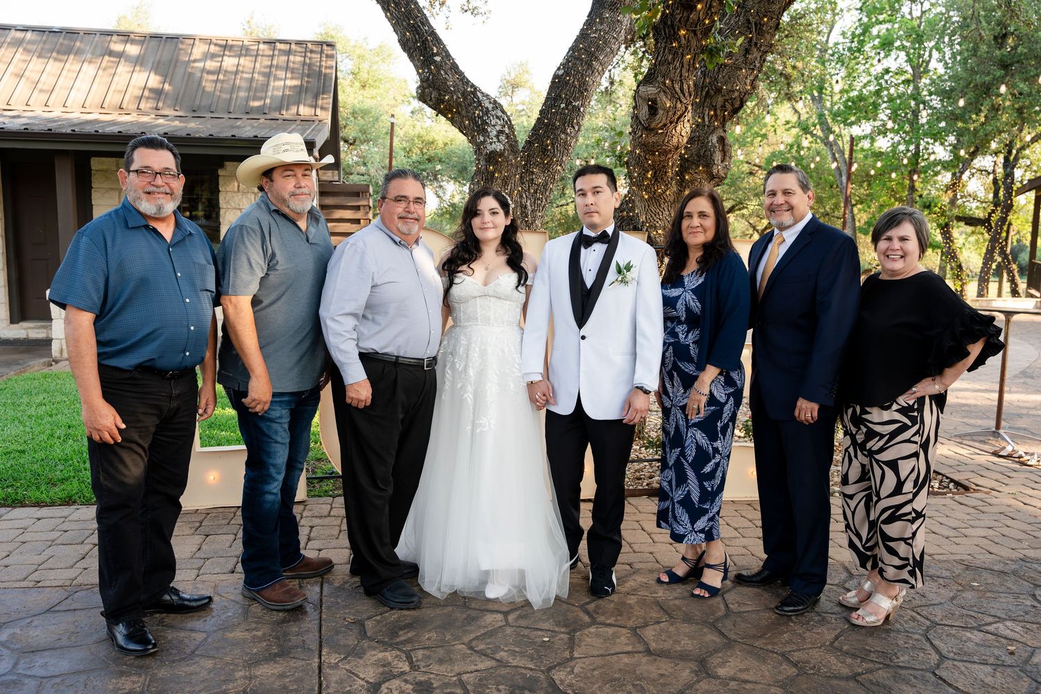 A wedding group poses together outdoors on a patio with trees and rustic building in the background.