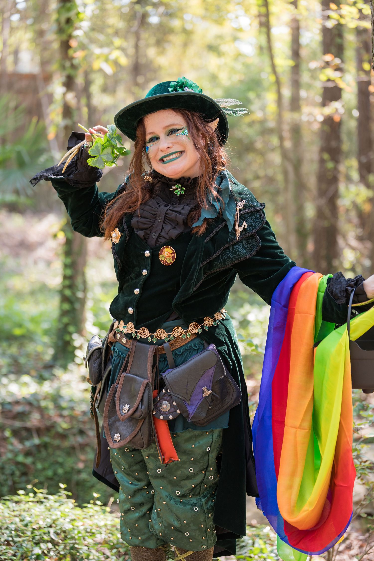 A costumed performer in green and black holds colorful ribbons in a forest setting.