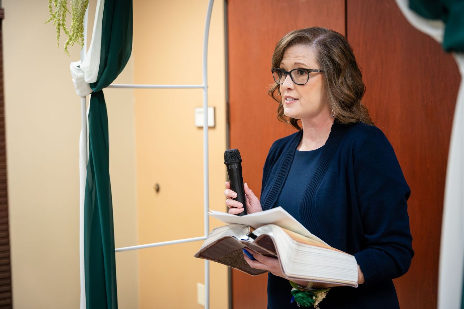 A person in a navy sweater holds a microphone and book while standing near emerald green curtains in a venue.