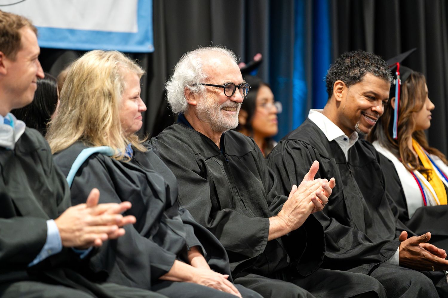 Row of attendees in academic regalia applauding during graduation ceremony.