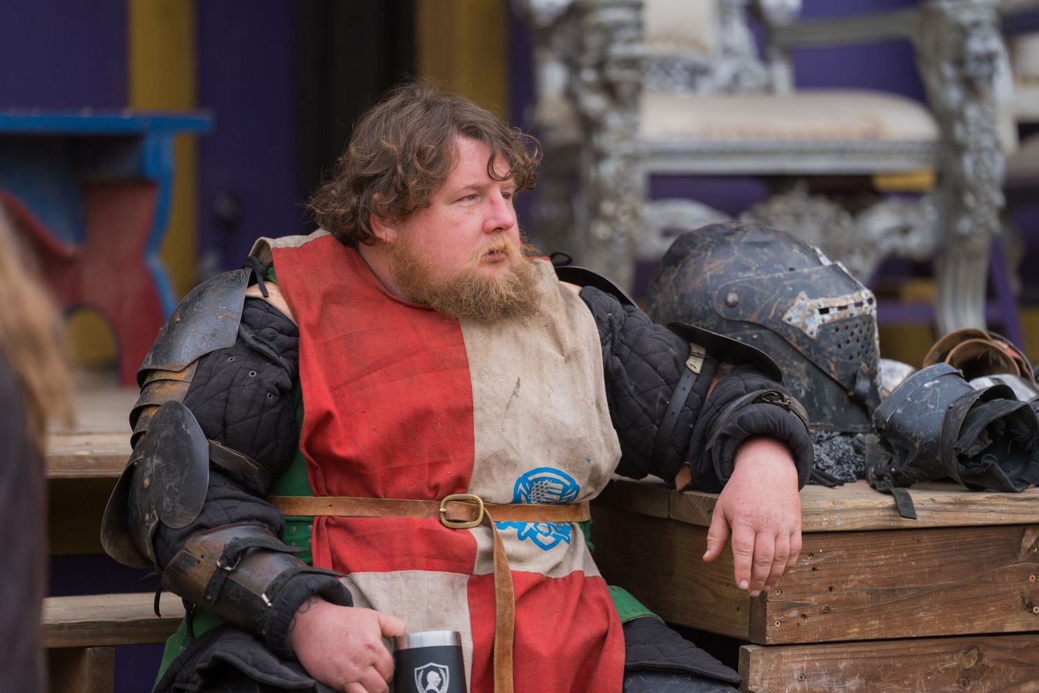 A performer in medieval armor and red tunic rests against wooden railings at a renaissance fair.