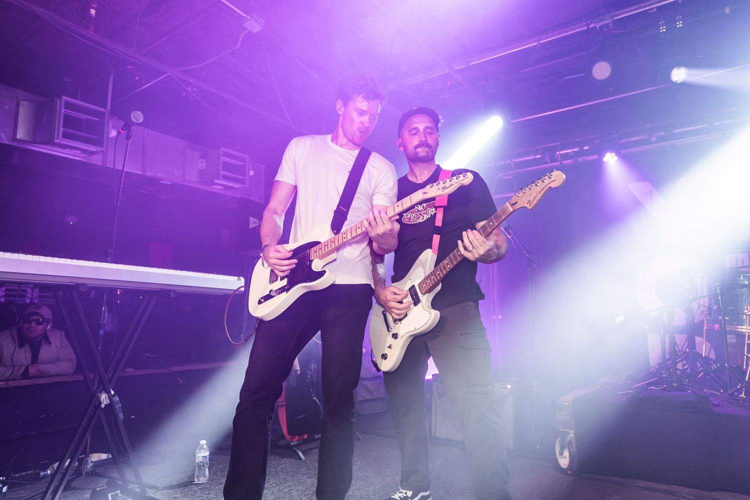 Musicians perform on stage with guitars under dramatic purple stage lighting and fog effects during a live concert.