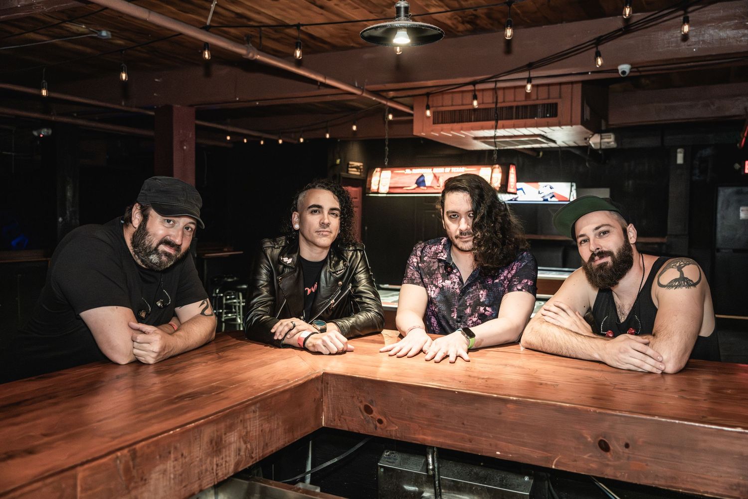 Rock band members seated at wooden table inside dimly lit venue.