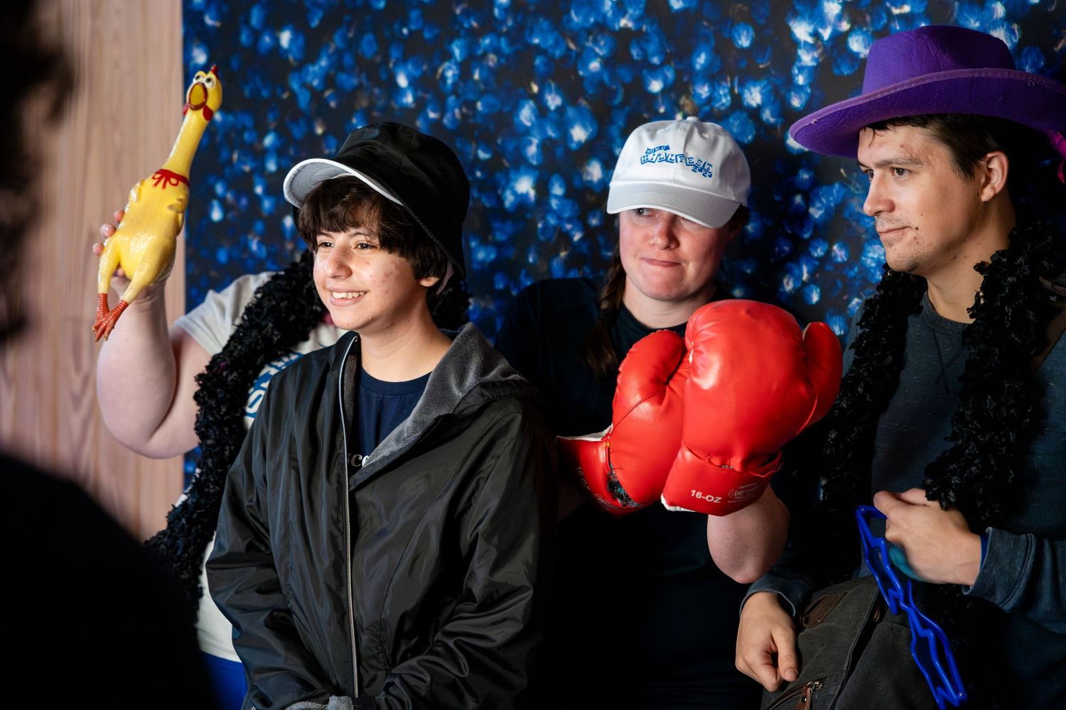 Three people posed together holding props including a yellow bottle and red boxing gloves against a blue sparkly background.
