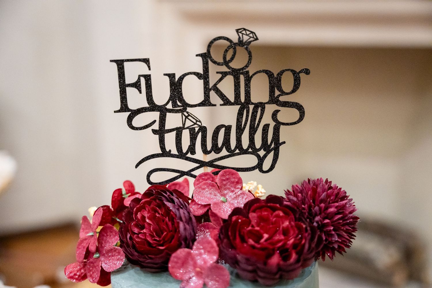 Wedding cake topper reading 'Fucking Finally' adorned with deep red and pink flowers.