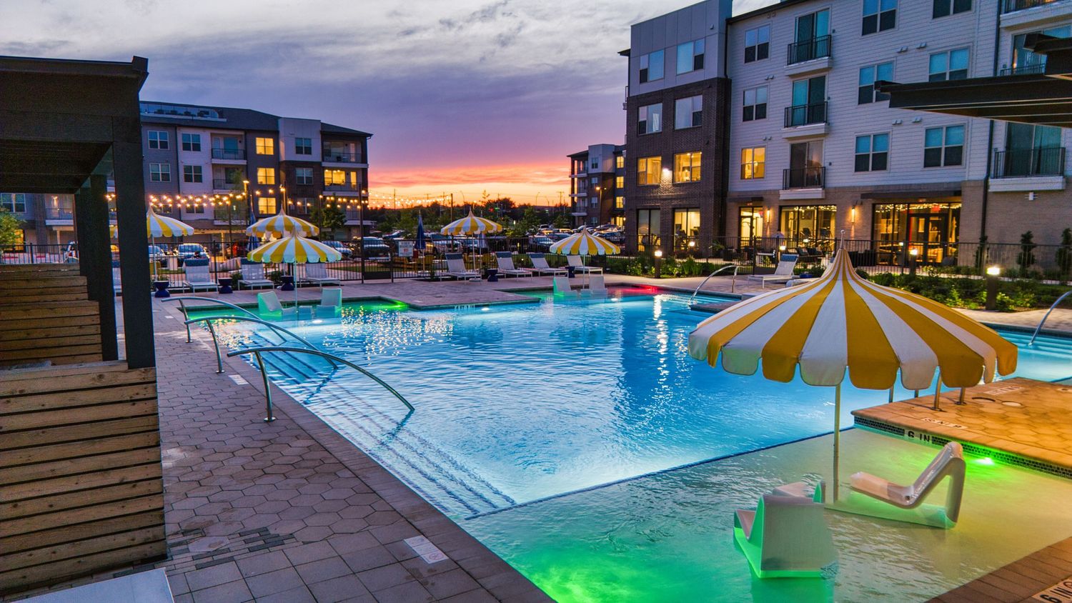 Illuminated pool area at dusk with striped umbrellas and modern apartment buildings.