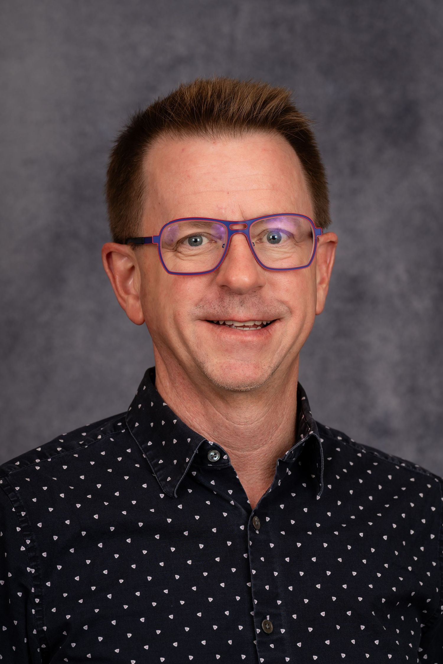 Professional headshot of person wearing purple glasses and polka dot shirt.