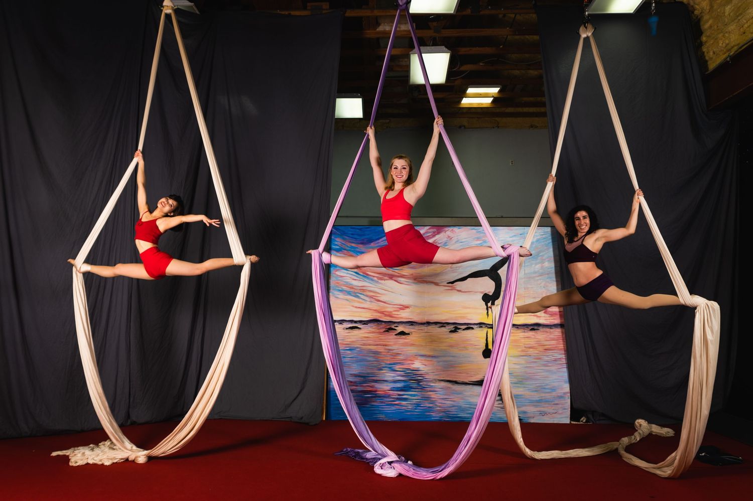 Three aerial artists perform synchronized poses in silks against a painted backdrop.